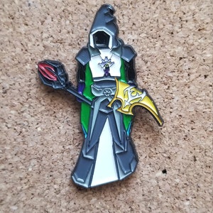 Puede incluir: Un pin de esmalte negro y verde que representa una figura encapuchada con una túnica blanca, sosteniendo un bastón rojo y negro y una guadaña dorada.