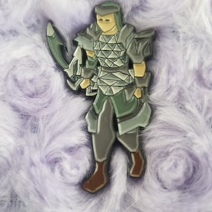 Puede incluir: Un pin de esmalte verde y gris de una figura de guerrero con una espada. El guerrero lleva armadura y casco. El pin está sobre un fondo difuso morado.