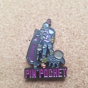 May include: A purple and black enamel pin featuring a knight in armor holding a bag of coins. The knight is standing on a black creature with a purple cape. The text "PIN POCKET" is written in purple below the image.