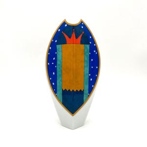 May include: A tall, pointed ceramic vase with a white base and a central shield-shaped design. The design features a red crown atop a large gold rectangle, flanked by teal panels, all set against a dark blue background with white speckles.