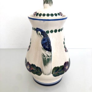 May include: White ceramic pitcher with a blue and green floral design. The pitcher has a lid with a green knob and a handle with a blue and green design.