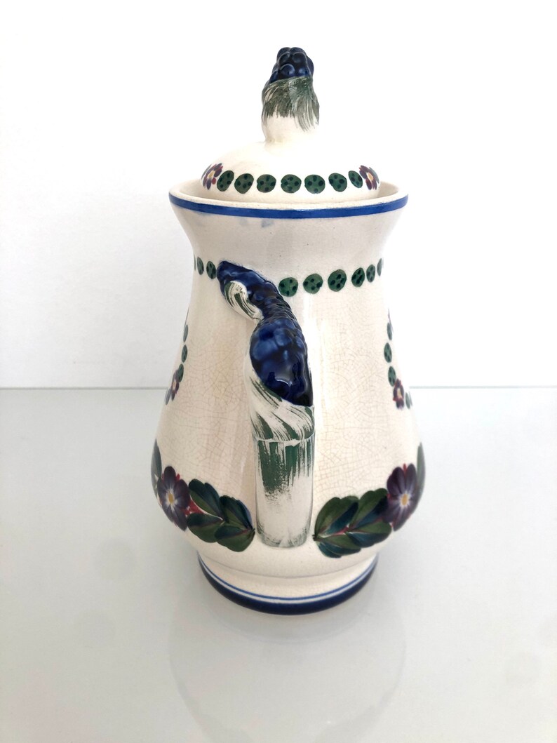 May include: White ceramic pitcher with a blue and green floral design. The pitcher has a lid with a green knob and a handle with a blue and green design.
