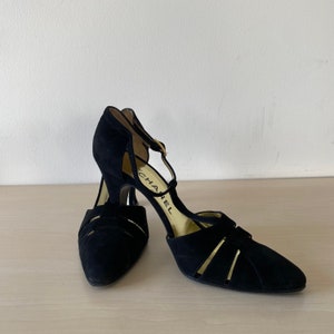Könnte beinhalten: Ein Paar schwarze Wildleder-High-Heels mit T-Riemen-Design. Die Schuhe haben eine spitze Zehenpartie und einen goldenen Schnallenverschluss. Der Markenname "CHANEL" ist auf der Innensohle sichtbar. Die Absätze sind etwa 7.6 cm hoch.