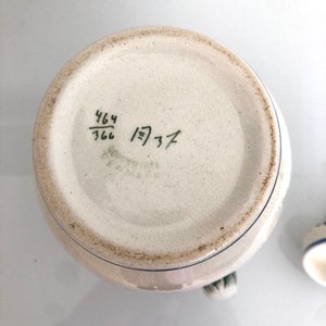 May include: The bottom of a white ceramic pitcher with blue and green accents. The bottom of the pitcher has a handwritten inscription that reads "464/766 1)37".