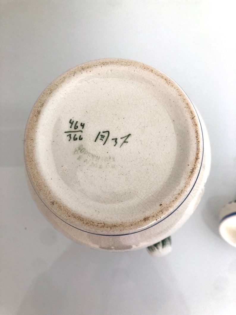 May include: The bottom of a white ceramic pitcher with blue and green accents. The bottom of the pitcher has a handwritten inscription that reads "464/766 1)37".