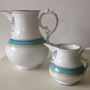 May include: A white ceramic pitcher and creamer set with a teal and gold trim. The pitcher has a curved handle and a spout. The creamer has a small handle and a spout.