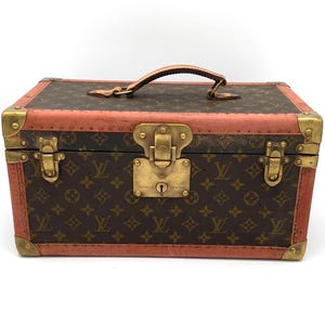 Może przedstawiać: Antyczna walizka podróżna Louis Vuitton z brązowym i beżowym wzorem monogramu. Walizka ma skórzany uchwyt i mosiężne okucia. Walizka jest zamknięta i ma czerwoną skórzaną obwódkę.
