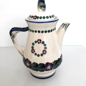 May include: A white ceramic teapot with a floral design. The teapot has a lid with a blue and green berry on top. The teapot has a blue and white trim around the base.