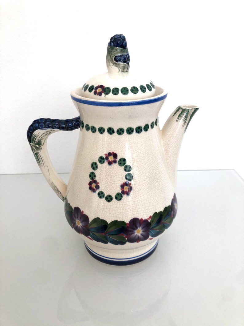 May include: A white ceramic teapot with a floral design. The teapot has a lid with a blue and green berry on top. The teapot has a blue and white trim around the base.