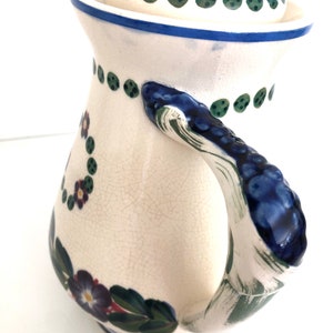 May include: A white ceramic pitcher with a blue and green floral design. The pitcher has a lid and a handle.