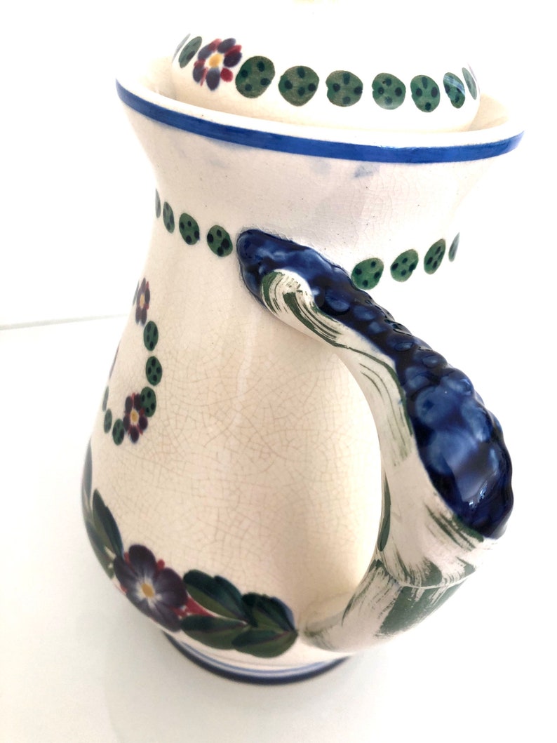 May include: A white ceramic pitcher with a blue and green floral design. The pitcher has a lid and a handle.