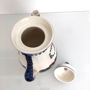 May include: A white ceramic teapot with a blue and green floral design. The teapot has a lid with a small hole in the top and a handle shaped like a bunch of grapes. The teapot is sitting on a white surface.