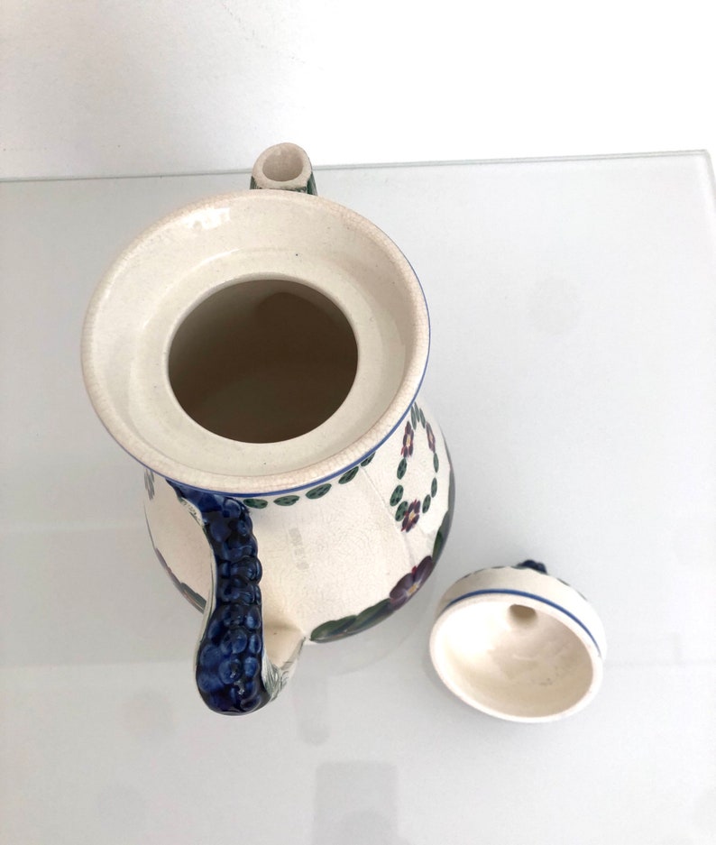 May include: A white ceramic teapot with a blue and green floral design. The teapot has a lid with a small hole in the top and a handle shaped like a bunch of grapes. The teapot is sitting on a white surface.