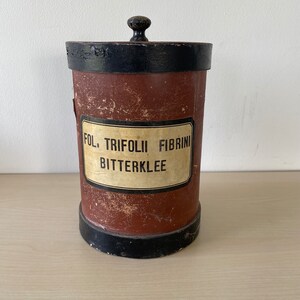 Può includere: Un contenitore vintage cilindrico in latta marrone e nero con un coperchio in legno e un'etichetta che recita "FOL. TRIFOLII FIBRINI BITTERKLEE".