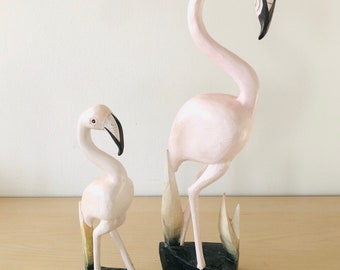 Puede incluir: Dos figuritas de flamenco de madera, una más grande que la otra, ambas pintadas de rosa con detalles negros. El flamenco más grande tiene la cabeza echada hacia atrás, mientras que el flamenco más pequeño tiene la cabeza girada hacia un lado. Ambos están de pie sobre bases negras con follaje pintado de verde.