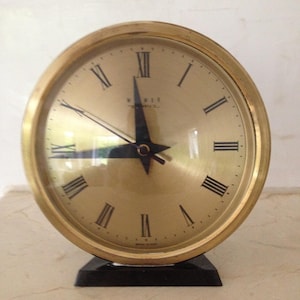 May include: A gold-colored analog clock with black Roman numeral markings on a white face. The clock has a black hour and minute hand and a black second hand. The clock is made in Japan and is sitting on a black base.