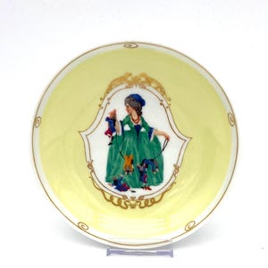May include: A decorative plate with a pale yellow background and a gold rim. The centre features a framed illustration of a person in a green dress holding puppets. The plate has a gold decorative border.