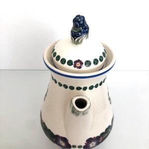 May include: White ceramic teapot with a floral design. The teapot has a lid with a blue and green berry design. The teapot has a spout and a handle.