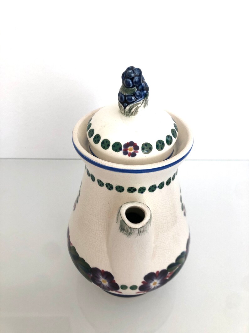 May include: White ceramic teapot with a floral design. The teapot has a lid with a blue and green berry design. The teapot has a spout and a handle.