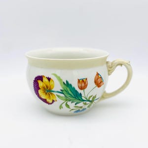 Puede incluir: Una taza de té de cerámica blanca con un diseño floral. La taza presenta una flor amarilla y morada, capullos naranjas y hojas verdes. El borde y el asa son de color amarillo claro.