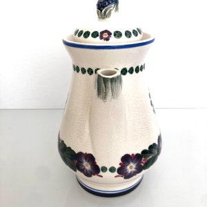 May include: White ceramic teapot with a floral design. The teapot has a lid with a grape cluster finial. The design features purple flowers, green leaves, and blue and green dots.