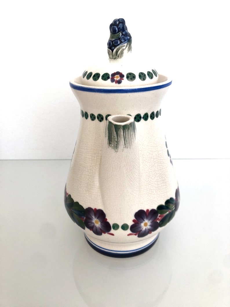 May include: White ceramic teapot with a floral design. The teapot has a lid with a grape cluster finial. The design features purple flowers, green leaves, and blue and green dots.