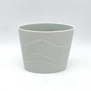 May include: A white ceramic planter with a textured, fan-like design on the lower half. The planter has a cylindrical shape and a smooth, matte finish. The design is a lighter shade of white, creating a subtle contrast.