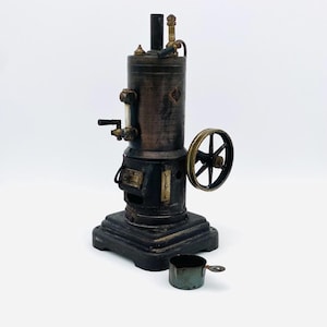 Könnte beinhalten: Ein Vintage-Dampfmaschinenmodell aus Messing und Schwarz mit einer kleinen, grünen Metalltasse. Die Maschine hat einen hohen, zylindrischen Kessel mit einem kleinen Rad und einem Rohr an der Seite. Das Modell steht auf einem schwarzen Sockel.