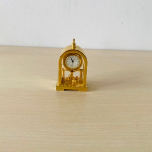 Puede incluir: Un reloj de abuelo en miniatura de tono dorado con una esfera blanca y agujas negras. El reloj tiene un diseño decorativo con un pequeño péndulo.