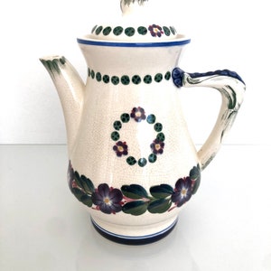 May include: A white ceramic teapot with a floral design. The teapot has a lid with a blue and green floral design. The teapot has a handle with a green and blue floral design.