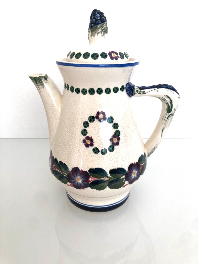 May include: A white ceramic teapot with a floral design. The teapot has a lid with a blue and green floral design. The teapot has a handle with a green and blue floral design.
