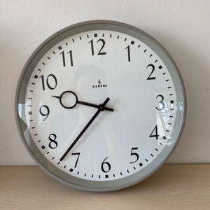 May include: A round wall clock with a white face, black numerals, and black hands. The clock has a gray bezel and the brand name "SIEMENS" is printed in black. The clock is on a light-colored surface.