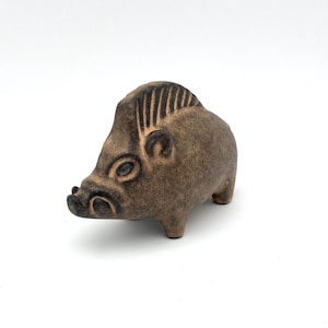 Puede incluir: Una figurita de cerámica marrón de un jabalí con ojos negros y una espalda texturizada. La figurita está de pie sobre una superficie blanca.