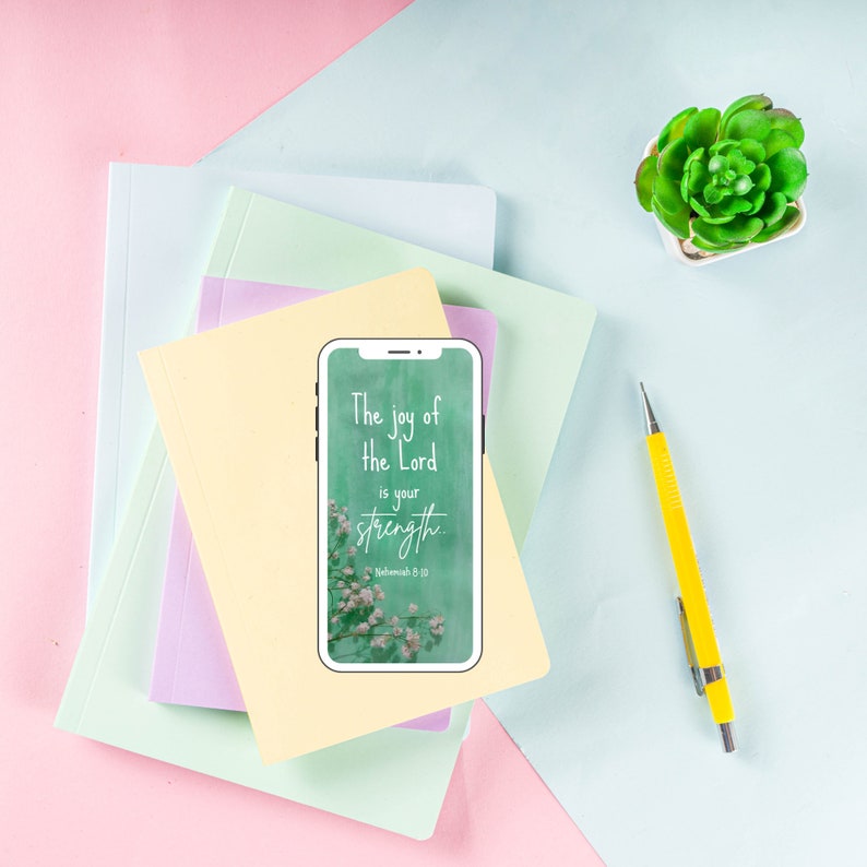 K&ouml;nnte beinhalten: Ein Smartphone zeigt einen gr&uuml;nen Hintergrund mit wei&szlig;en Blumen und den Text "The joy of the Lord is your strength... Nehemiah 8:10". Das Telefon liegt auf einem Stapel bunter Notizb&uuml;cher.