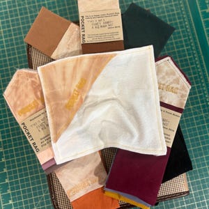 May include: A collection of organic cotton pocket rags in various colors, including white, orange, and burgundy. Each rag has a unique design and is labeled with text that reads "THIS IS NOT A FANCY HANKY - A KID MADE THIS!"