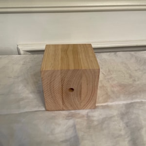 May include: A small, square, wooden block with a hole in the center. The block is made of light-colored wood with visible grain.
