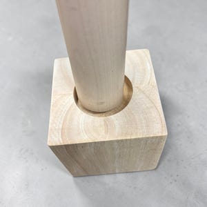 May include: A light-colored wooden object consisting of a vertical cylindrical post securely fitted into a square block base. The natural wood grain is visible throughout the structure, showing a smooth, unfinished surface texture on both parts.