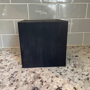 May include: A black wooden square block, possibly a display stand or riser, sitting on a speckled countertop.