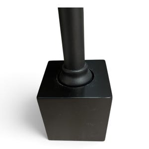 May include: A matte black square base with a cylindrical post extending upward from the center. The base features a smooth, solid construction with a rounded collar where the vertical post connects to the top surface of the square block.