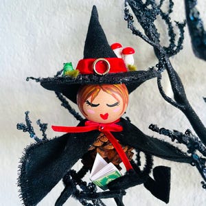 May include: A decorative figurine featuring a wooden head with painted facial features, wearing a black felt witch hat with a red ribbon, a gold ring, and small mushroom accents. The body is a pinecone with a black felt cape and a tiny green book.