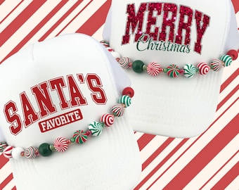 May include: Two white trucker hats with red text that reads "SANTA'S FAVORITE" and "MERRY Christmas". Each hat has a beaded garland of red, green, and white candy-themed beads. The background has a red and white candy cane stripe pattern.