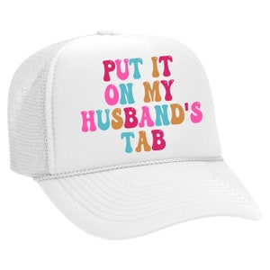May include: White trucker hat with a mesh back. The hat has the text "PUT IT ON MY HUSBAND'S TAB" in colorful, retro-style letters.