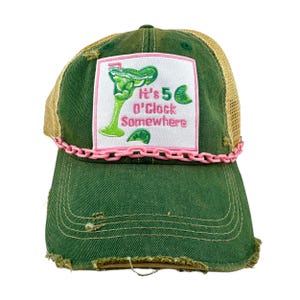 May include: Green and brown trucker hat with a pink chain trim. The hat has a white patch with a green margarita glass and the text "It's 5 O'Clock Somewhere" in pink.