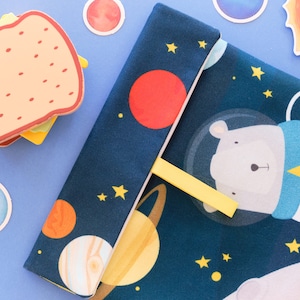 Può includere: Una copertina di libro in tessuto blu navy con un design di orso polare astronauta, pianeti e stelle. È inserito un segnalibro giallo. Adesivi decorativi a forma di sandwich e pianeti sono sparsi.
