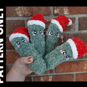 May include: Four green crocheted plush toys shaped like cucumbers with white eyes and red smiling mouths. Each toy is wearing a red and white crocheted Santa hat.