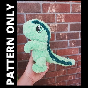 May include: A green crocheted dinosaur plush toy with a dark green stripe down its back and a single large eye. The toy is standing on a hand.