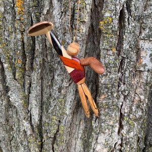 May include: A wooden figurine of a person holding a disc, carved from different types of wood, with a red, black, and white striped body and a brown head. The figurine is hanging from a string.