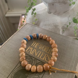 Peut inclure: Un bracelet de perles composé de perles en bois brun clair et de perles bleues. Le bracelet est posé sur un livre avec des lettres dorées qui dit "THE WORLD'S PAINTERS". Le bracelet est noué avec une ficelle de couleur naturelle.