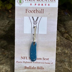 Könnte beinhalten: Ein blauer, fußballförmiger Anhänger aus einem Kunststadiumsitz, befestigt an einem Metall-Reißverschluss. Der Anhänger befindet sich auf einer Karte mit dem Text "Football" und "NFL Stadium Seat" und "Buffalo Bills".