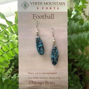 Op de afbeelding: Ovale oorbellen met een turquoise en zwart gespikkeld ontwerp, opgehangen aan zilverkleurige haken. De kaart achter de oorbellen leest "Football" en vermeldt de Chicago Bears.